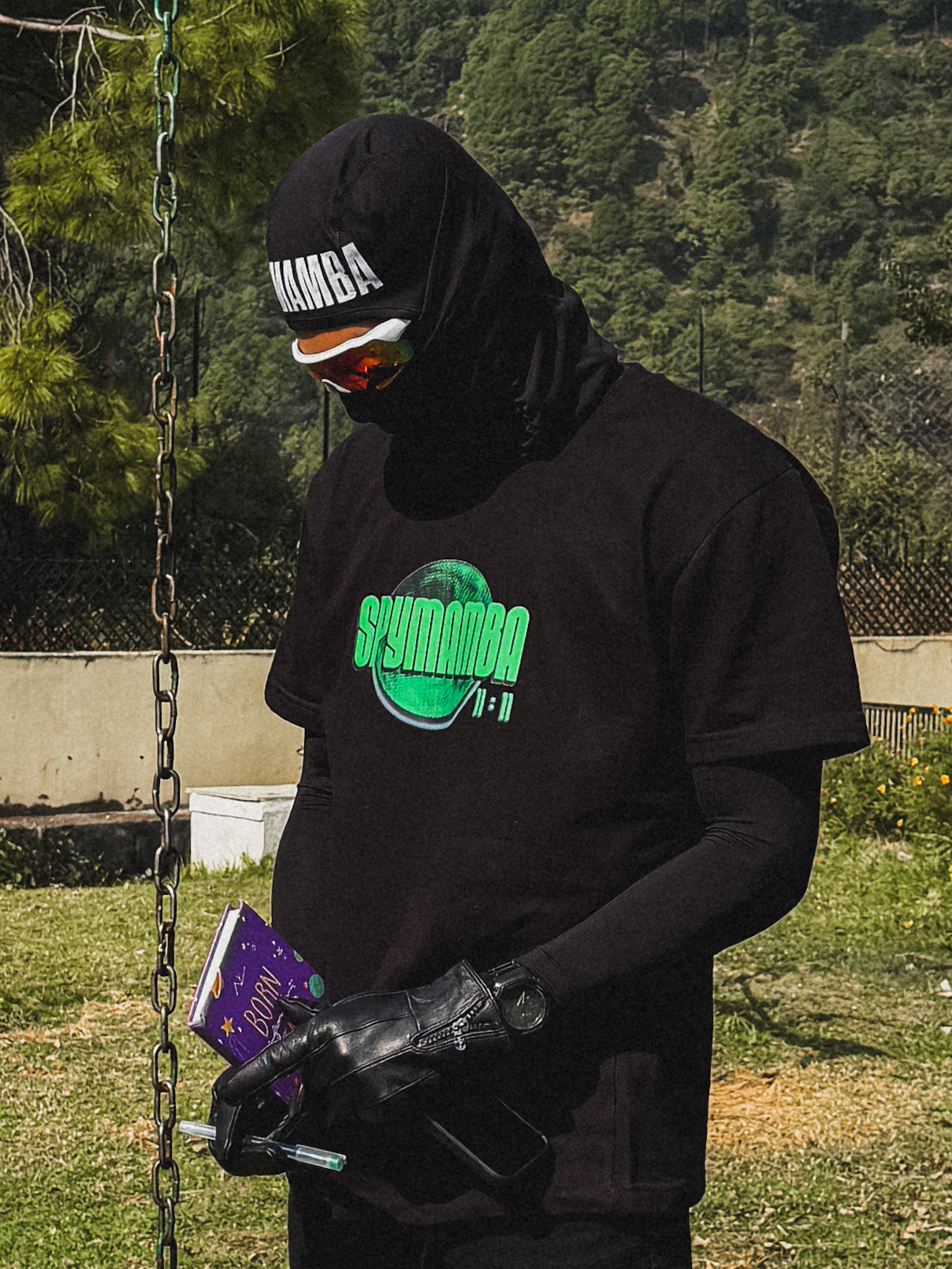 Person wearing a black hoodie and gloves, holding a book, with a forest background.