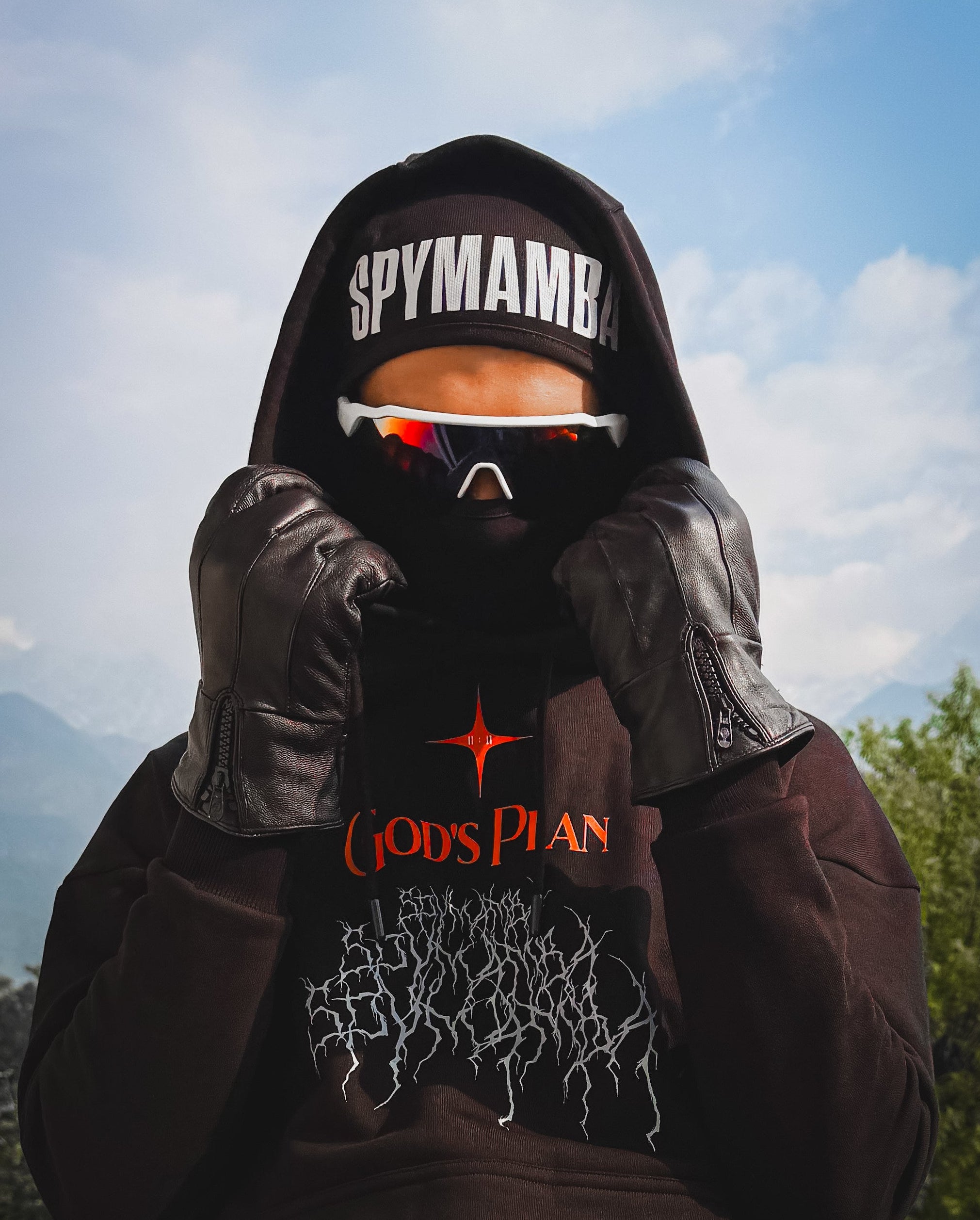 Person wearing a hoodie with 'SPYMAMBA' text, sunglasses, and leather gloves outdoors.